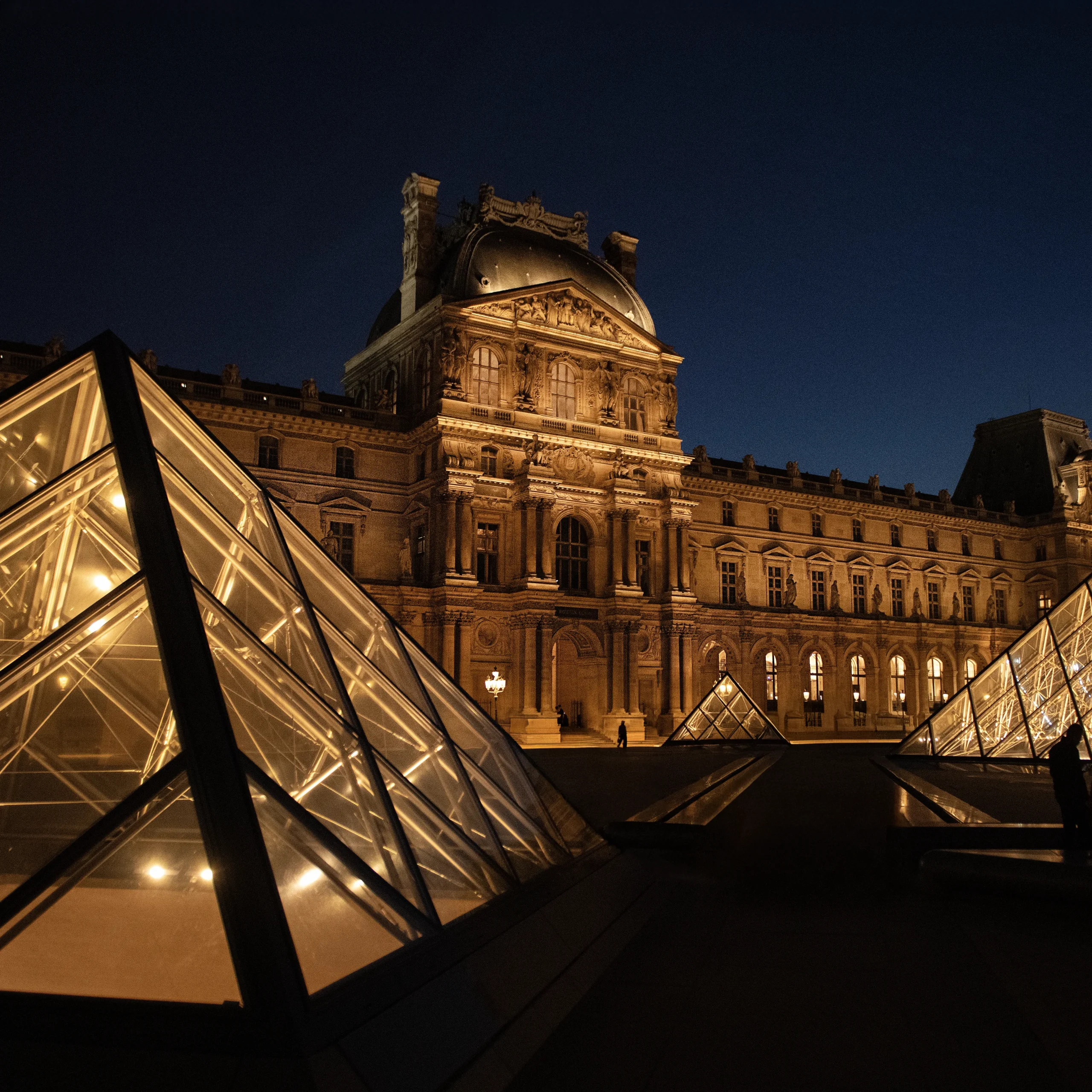Szklana piramida Luwru nocą w Paryżu, oświetlona złotym światłem na tle historycznej fasady muzeum